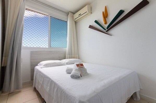 A hotel room with a bed, a window with curtains, an air conditioning unit, and a wall-mounted shelf with decorative items. - CJC Short Stay Blue Ocean (Natal)