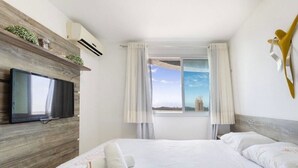 A hotel room with a bed, a mounted TV, a window with curtains, and a view of a building outside. - CJC Short Stay Blue Ocean (Natal)