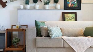 A living room with a beige sofa, a wooden side table, and a potted plant. - CJC Short Stay Blue Ocean (Natal)