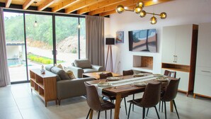 A spacious living area with a dining table, chairs, a sofa, and a large window offering a view of a garden and a road. - Nubária Residences l (San Pablo Etla)