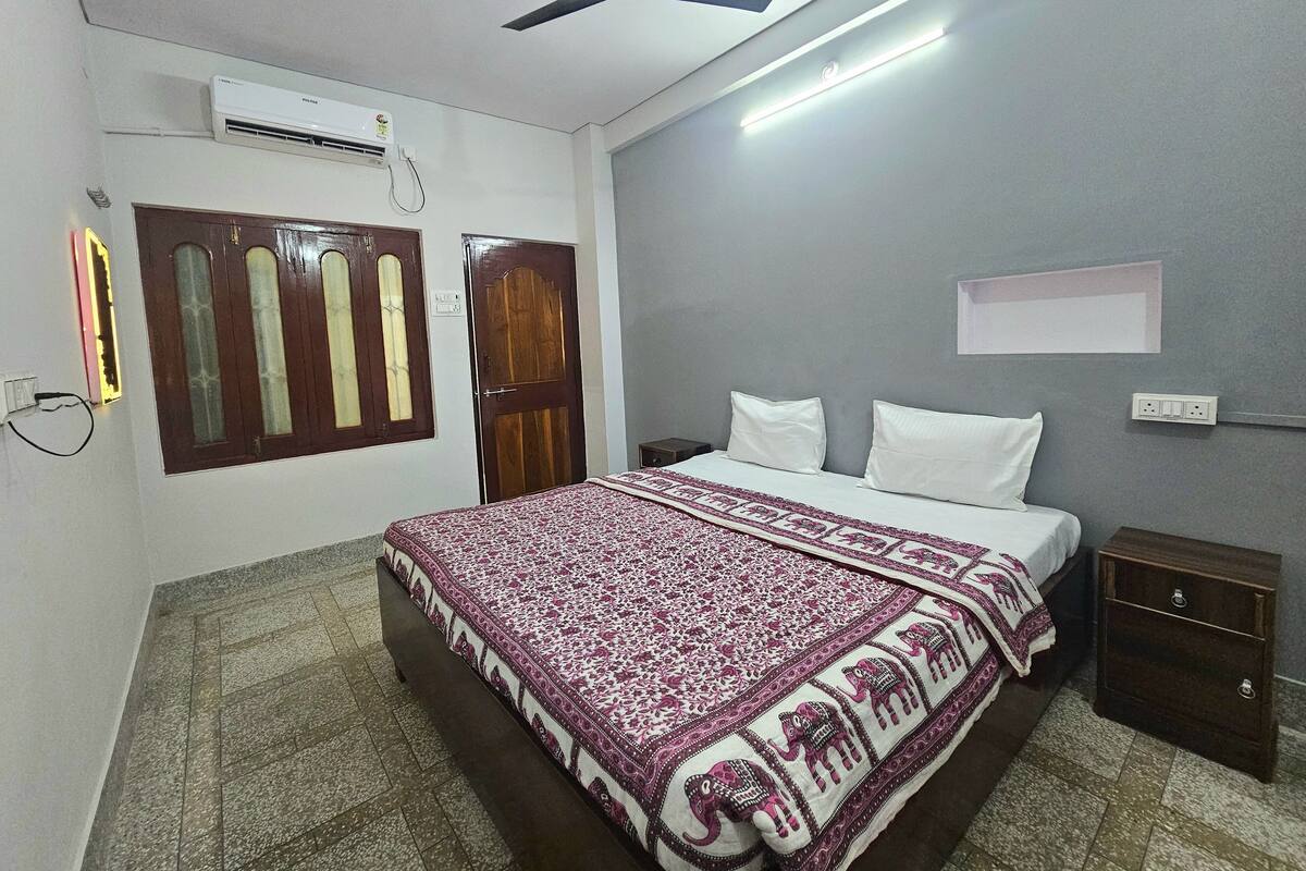 A bedroom with a bed, a nightstand, a ceiling fan, an air conditioning unit, and a window with a wooden door.
