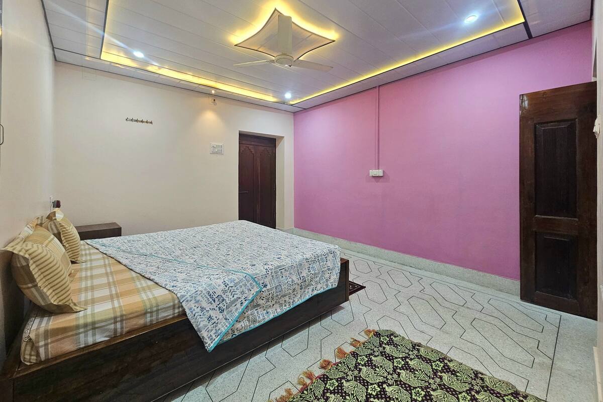 A bedroom with a bed, a wooden door, a pink wall, and a ceiling light fixture.