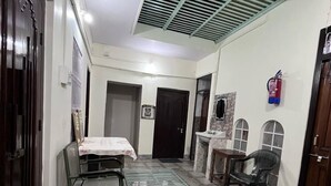 A hallway with a tiled floor, a metal grate, a table with chairs, a fire extinguisher, and a door. - Saraogi Homestays (Varanasi)