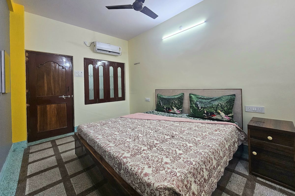 A bedroom with a bed, a ceiling fan, an air conditioner, and a nightstand.