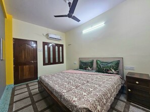 A bedroom with a bed, a ceiling fan, an air conditioner, and a nightstand. - Saraogi Homestays (Varanasi)