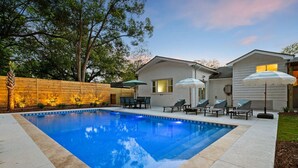 A rectangular swimming pool with lounge chairs and umbrellas, surrounded by a wooden fence and a house.