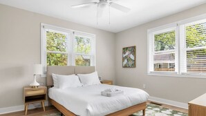 Un dormitorio con cama, mesitas de noche, lámpara, un cuadro y ventanas con persianas.