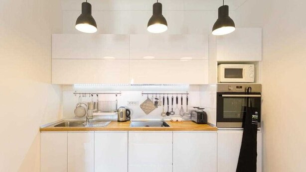 Una cocina moderna con armarios blancos, encimera de madera, un lavamanos, un hervidor, un tostador, un microondas y un horno. Hay tres lámparas colgantes sobre la encimera.