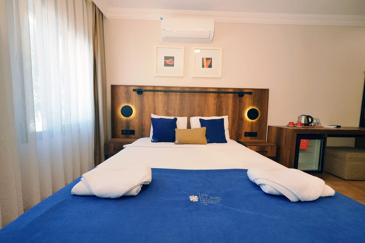 Una habitación de hotel con una cama grande, un cabecero de madera, dos lámparas fijadas a la pared, una mesita con hervidor de agua y un minibar.