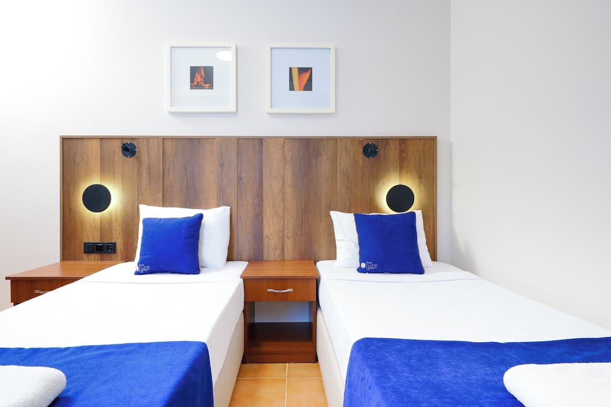 Habitación de hotel con dos camas, cabecera de madera, ropa de cama blanca, mantas de color azul y dos cuadros en la pared.