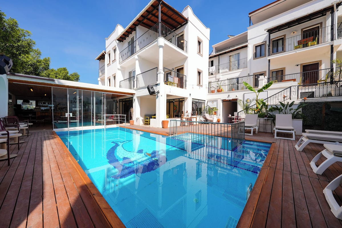 Zona de piscina con terraza de madera, bar con paredes de cristal y un edificio de varios pisos con balcones.