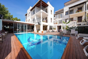 Uma área de piscina com um deque de madeira, um bar com paredes de vidro e um edifício de vários andares com varandas.