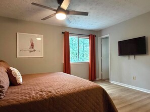 A bedroom with a bed, a ceiling fan, a window with curtains, a door, and a flat-screen TV mounted on the wall.