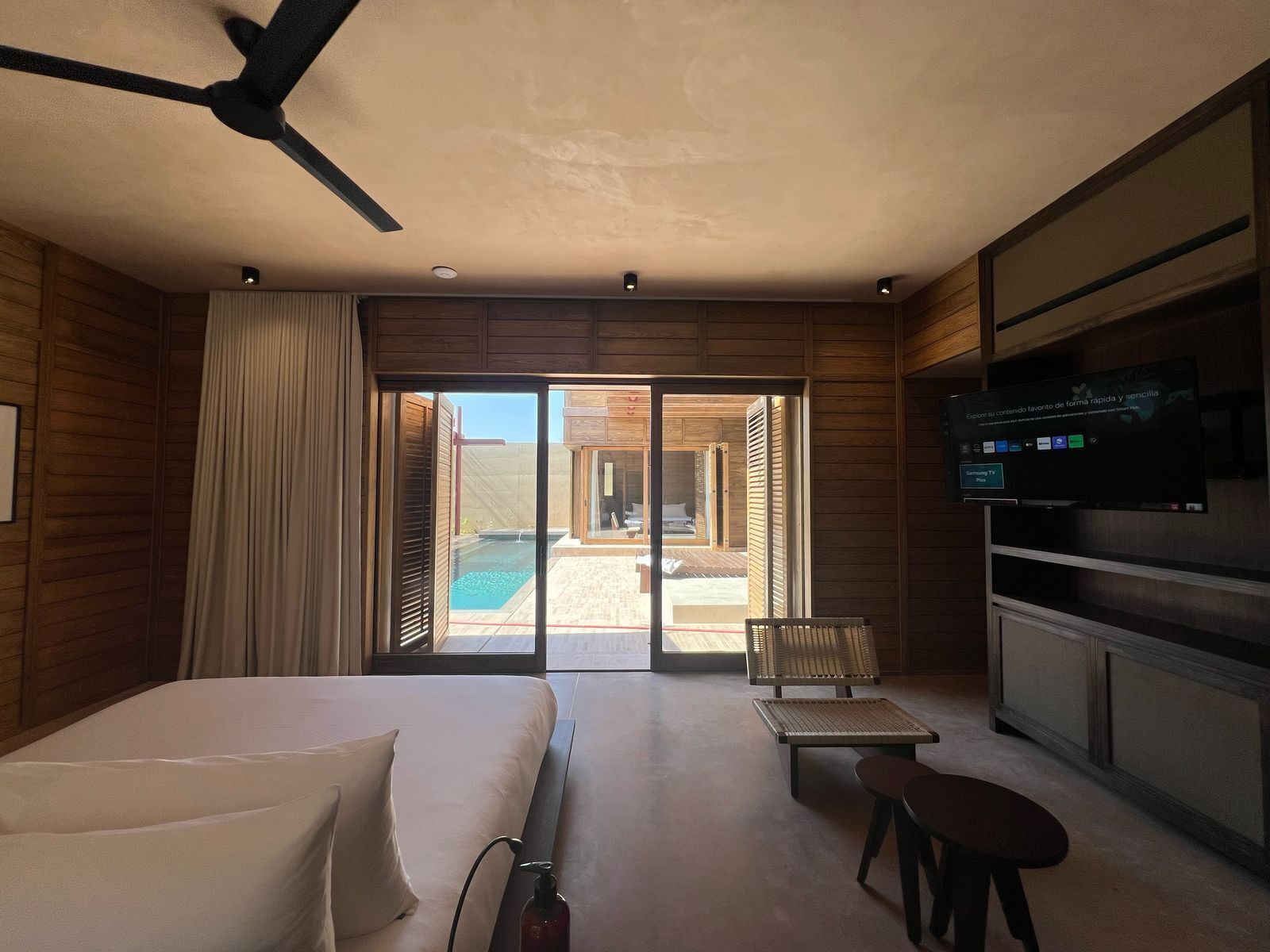 A hotel room with a bed, a TV, a ceiling fan, a sliding glass door to a pool area, and a small table with a chair.