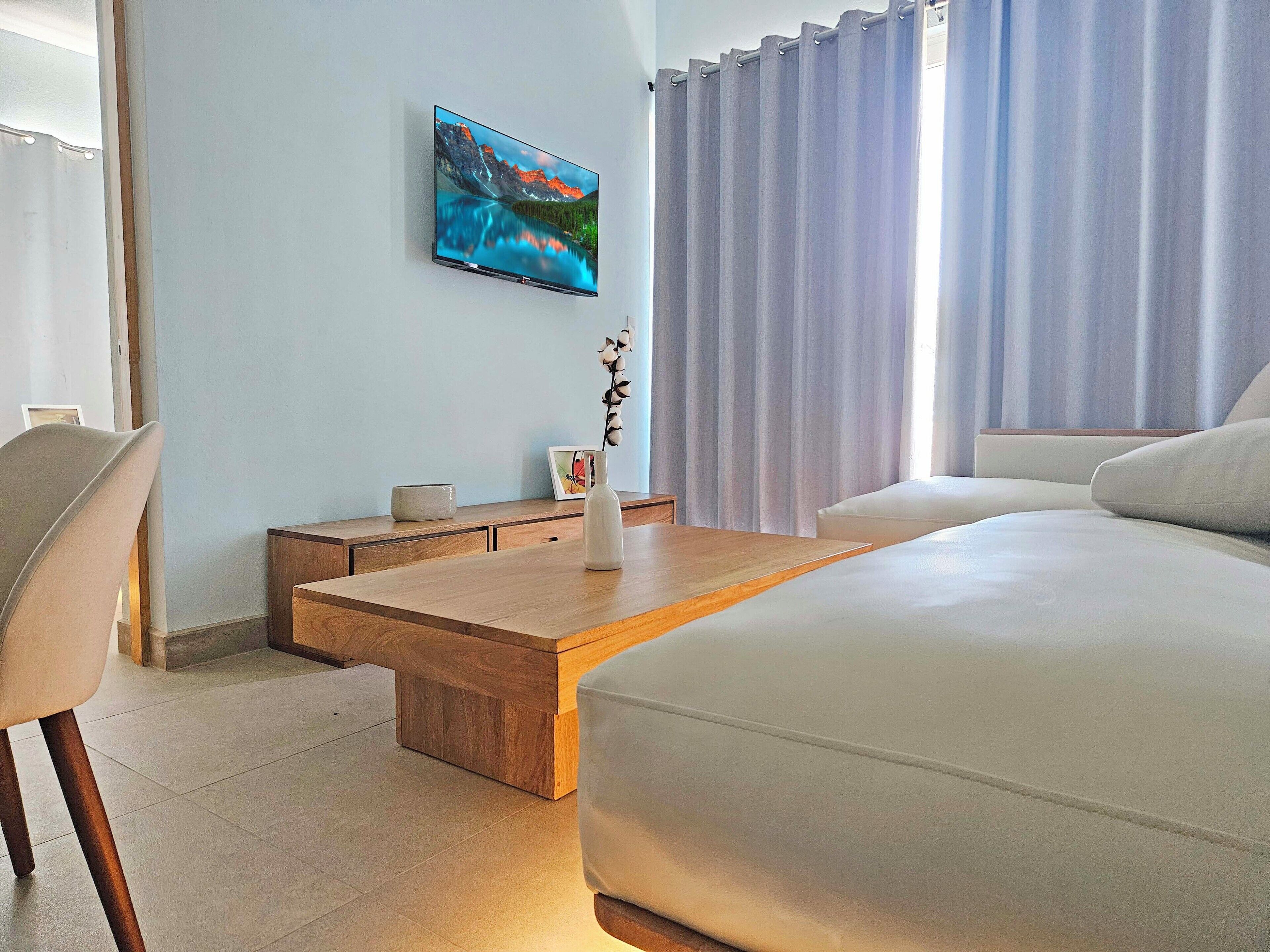 A modern hotel room with a large bed, a wooden coffee table, a TV mounted on the wall, and a view of mountains through the window.