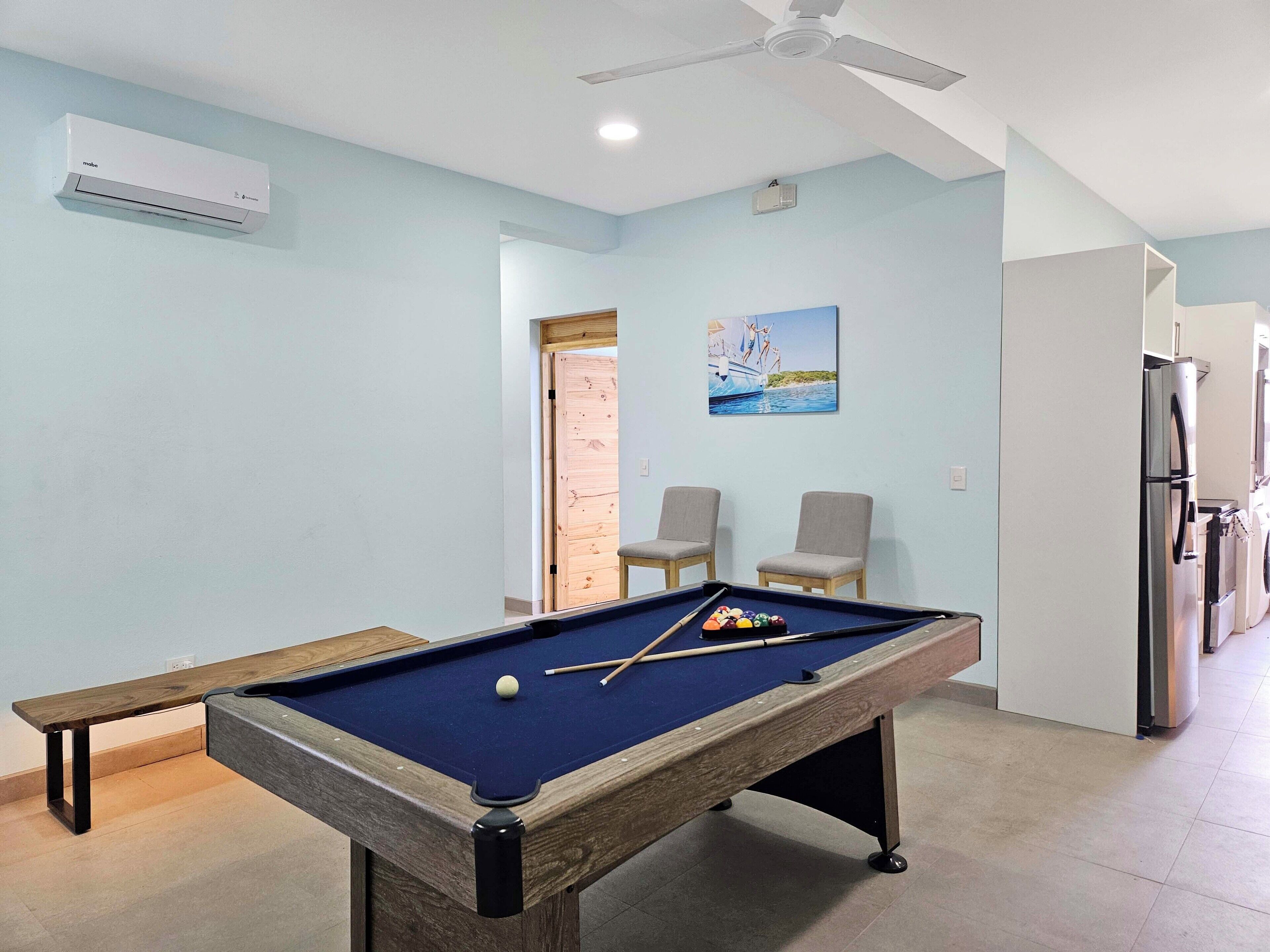 A pool table with a blue felt surface, a wooden bench, and a ceiling fan in a room with light blue walls.