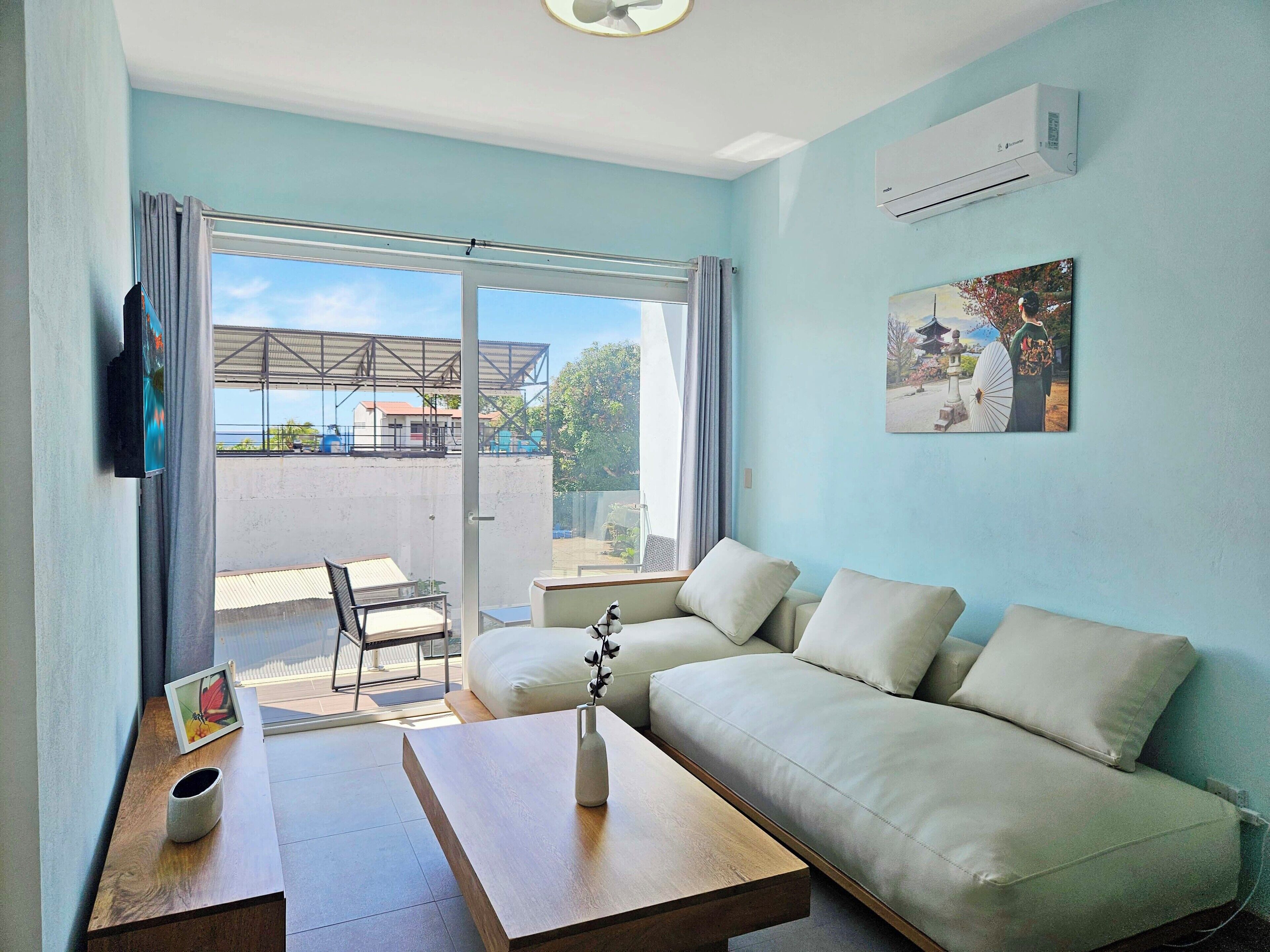 A modern living room with a sofa, a coffee table, and a TV. There is a large window with a view of a building and trees.
