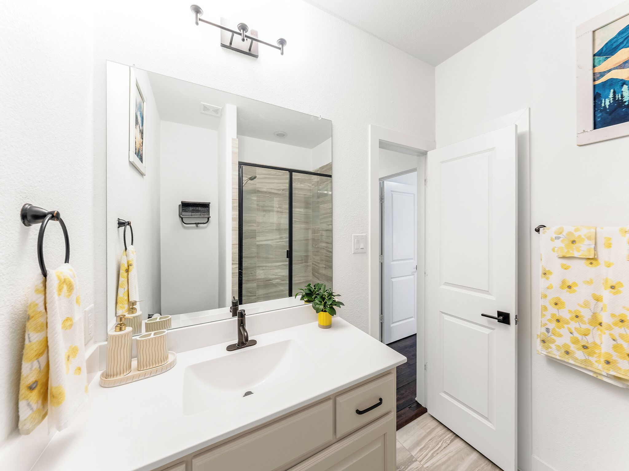 Un baño moderno con tocador blanco, un espejo grande, cabina de ducha con mampara de vidrio y una puerta que da a otra habitación.
