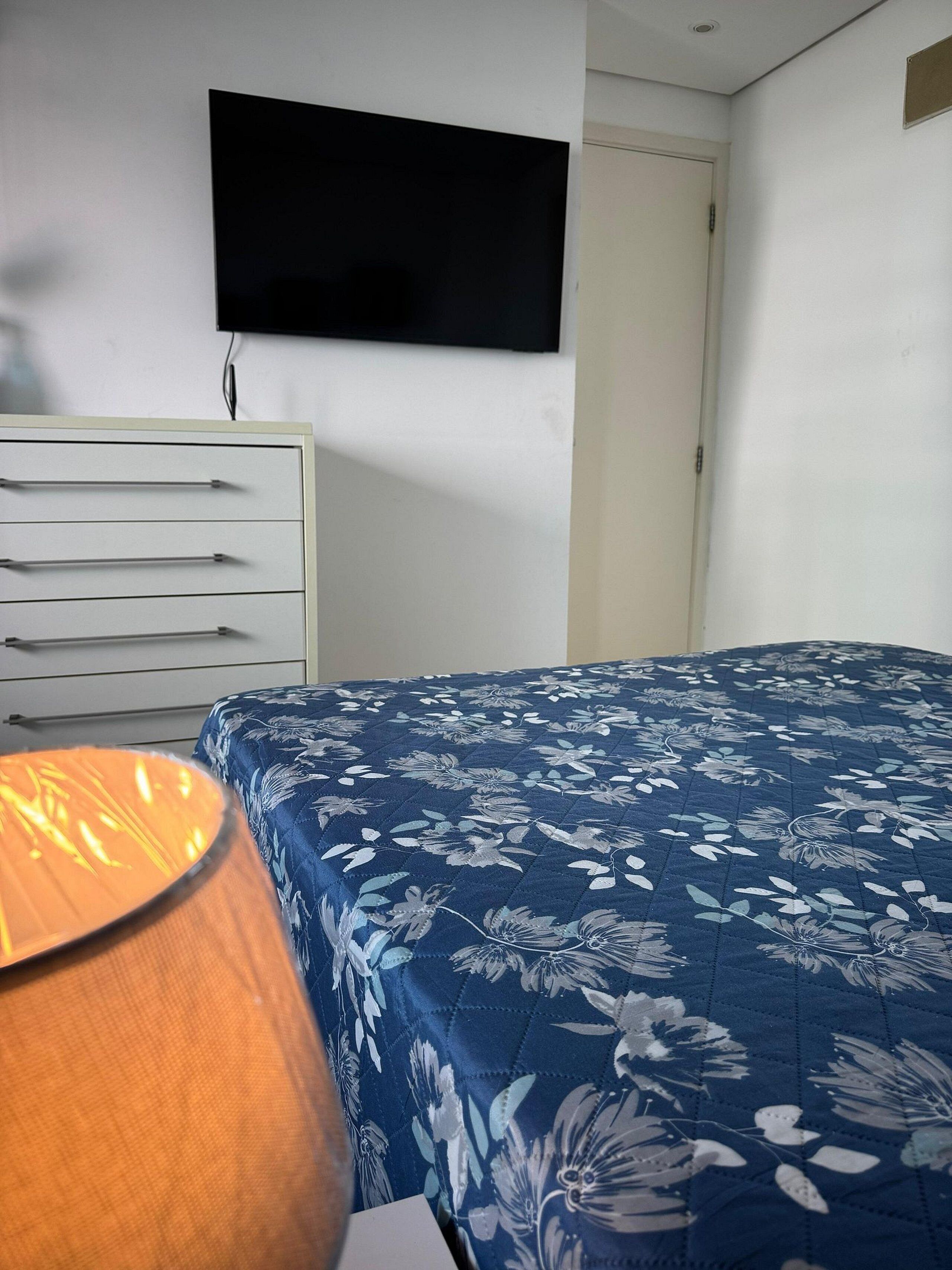 A bedroom with a bed, a dresser, a television on the wall, and a lamp.