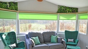 Une pièce avec un canapé gris, deux fauteuils verts et une vue sur un jardin, visible à travers de grandes fenêtres équipées de stores verts.