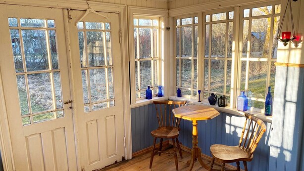 A room with a wooden floor, a table with chairs, and multiple windows with views of trees. - Charming Old Cottage in Scenic Southern Småland, Just Outside Markaryd | SE07057 (Markaryd)