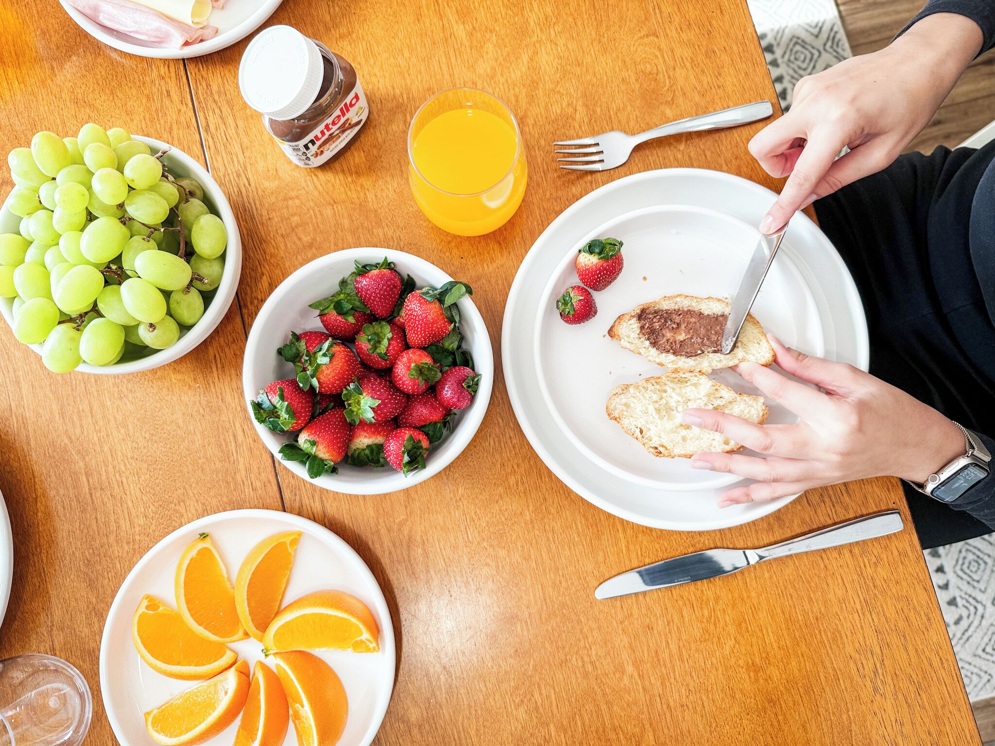 A table with a plate of toast with Nutella, a bowl of strawberries, a bowl of green grapes, a glass of orange juice, and a plate with orange slices.