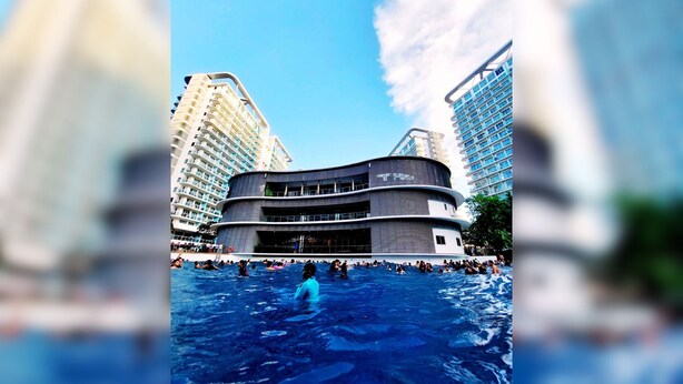 Un hotel moderno con fachada curva, varios balcones y una zona de piscina con gente nadando.