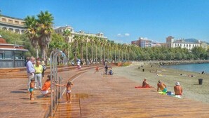 Una pasarela de madera con gente caminando y sentada, con vista a una playa con palmeras y edificios al fondo.