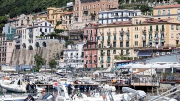 Un pueblo costero con edificios históricos, un puerto deportivo con barcos y una colina con vegetación.