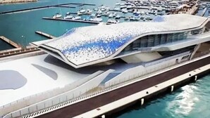 Un edificio moderno frente al agua con un techo curvo distintivo, un largo paseo peatonal y un puerto deportivo con barcos.