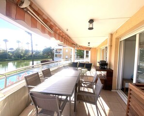 Une terrasse extérieure couverte, avec une table et des chaises, un barbecue, et une vue sur une étendue d’eau et des bâtiments en arrière-plan.