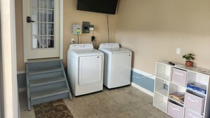 Une buanderie équipée de deux lave-linge et deux sèche-linge blancs, d’un téléviseur fixé au mur et d’une petite étagère avec des boîtes de rangement.