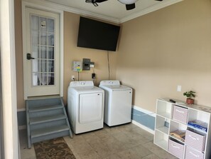 A laundry room with two white washers and dryers, a TV mounted on the wall, and a small shelving unit with storage boxes.