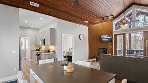 A modern kitchen with white cabinets, a dining table with chairs, and a living area with a gray sofa and a TV.