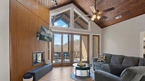 A spacious living room with a high ceiling, wooden paneling, a fireplace, a television, and a large sliding glass door leading to an outdoor patio. - Mountain Sanctuary, Balcony, View, and Grill! (Sylva)