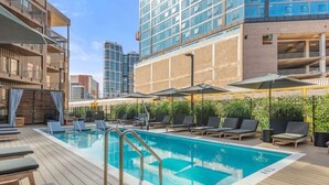 Una piscina en la azotea con sillones de descanso y sombrillas, rodeada de edificios altos.