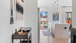 A modern kitchen with a central island, a dining area with a wooden table, and a view of a cityscape through large windows. - Cuddly Cactus for 12|Wraparound Balcony, Pool, Gym (Nashville)
