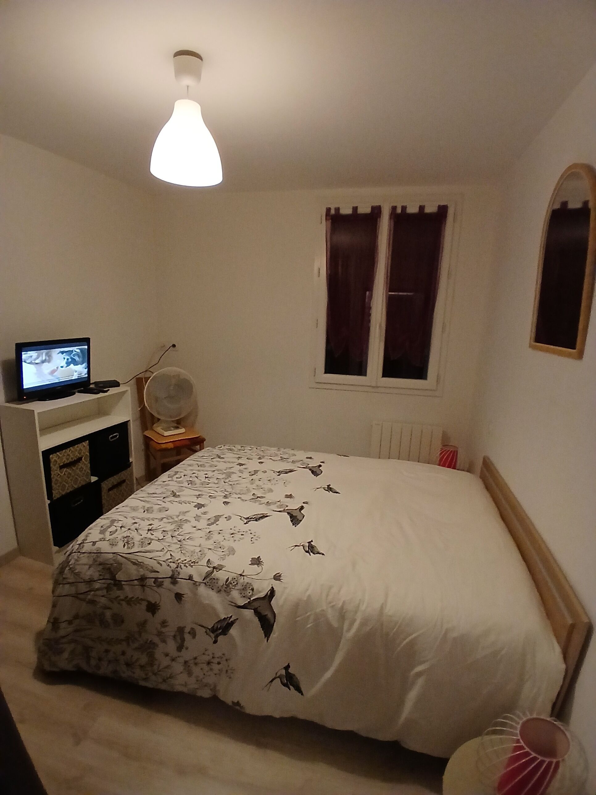 Une chambre à coucher avec un lit, un bureau avec un ordinateur, une fenêtre avec des rideaux et un radiateur.