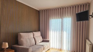 Una habitación de hotel moderna con sofá, televisor montado en la pared y un ventanal con cortinas.