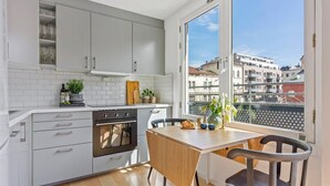 Une cuisine moderne avec des meubles blancs, une table en bois et une vue sur les bâtiments, visible à travers une grande fenêtre.