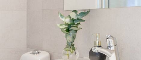 Ein Badezimmer mit einem weißen Waschbecken, einer Glasvase mit grünen Blättern, einer Seifenspender und einem Spiegel über dem Waschbecken.