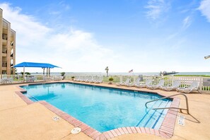 A swimming pool with lounge chairs and a blue umbrella, surrounded by a white fence, with a view of the ocean.