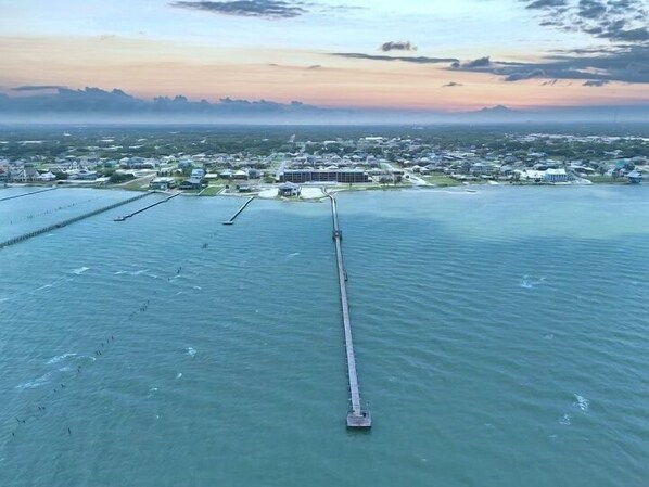 An aerial view of a coastal area with a long pier extending into the water, surrounded by residential buildings and a clear sky.