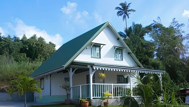 A two-story house with a green roof and white walls, surrounded by lush greenery.