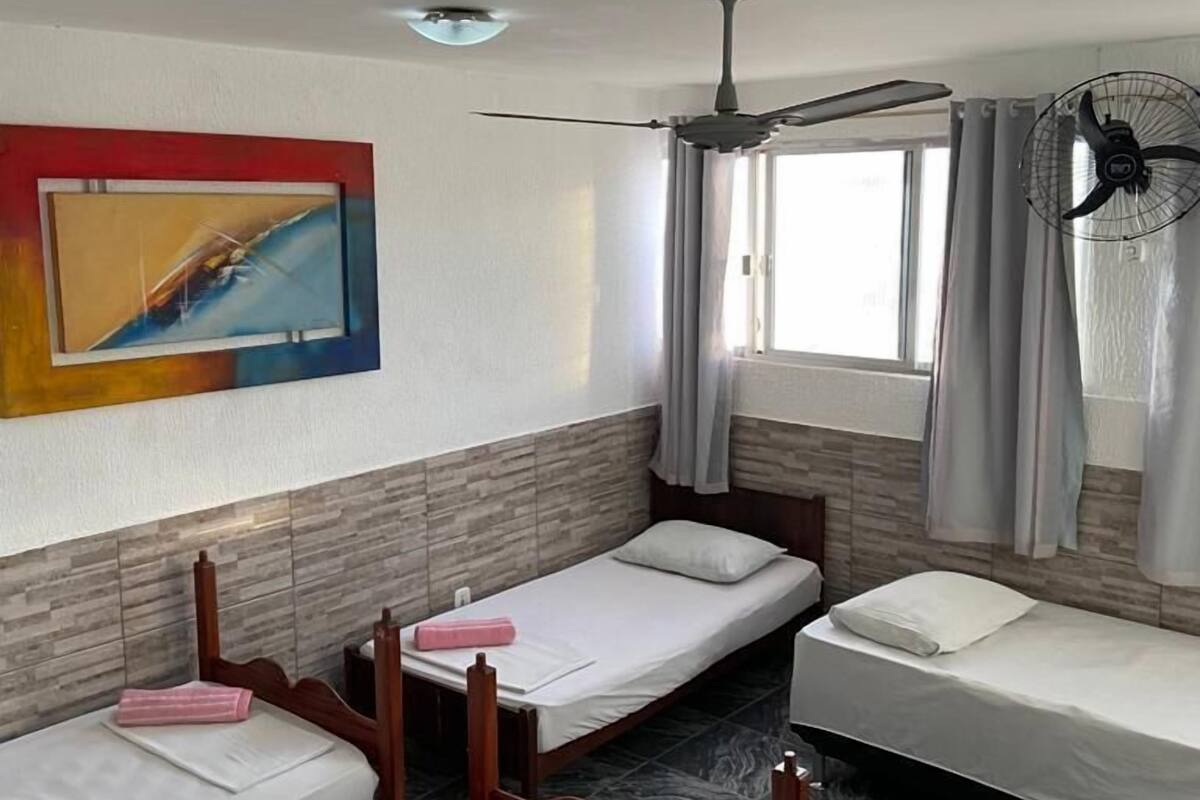 Una habitación con tres camas individuales, ventilador de techo, ventana con cortinas y ventilador de pared.