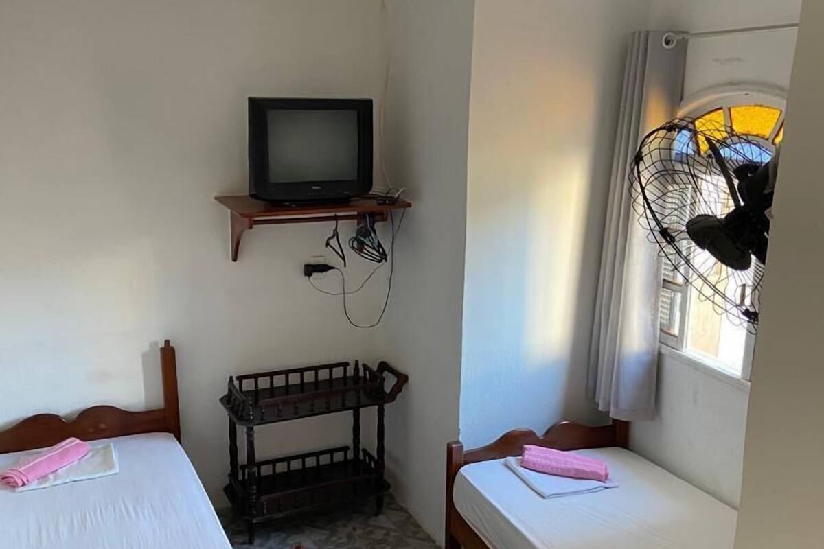 Una habitación pequeña y sencilla con dos camas, un televisor y una ventana con ventilador.