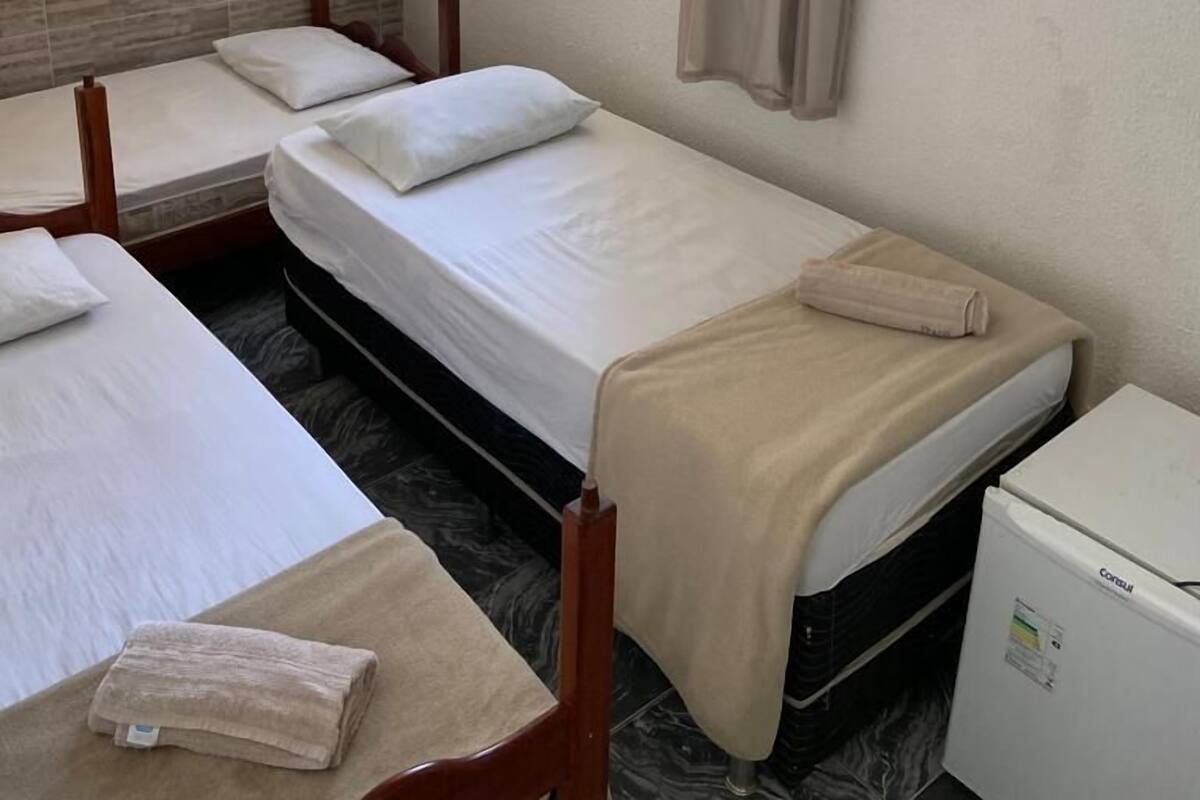 Habitación de hotel con tres camas individuales, ventilador de techo, ventana con cortinas, refrigerador pequeño y teléfono fijo en la pared.