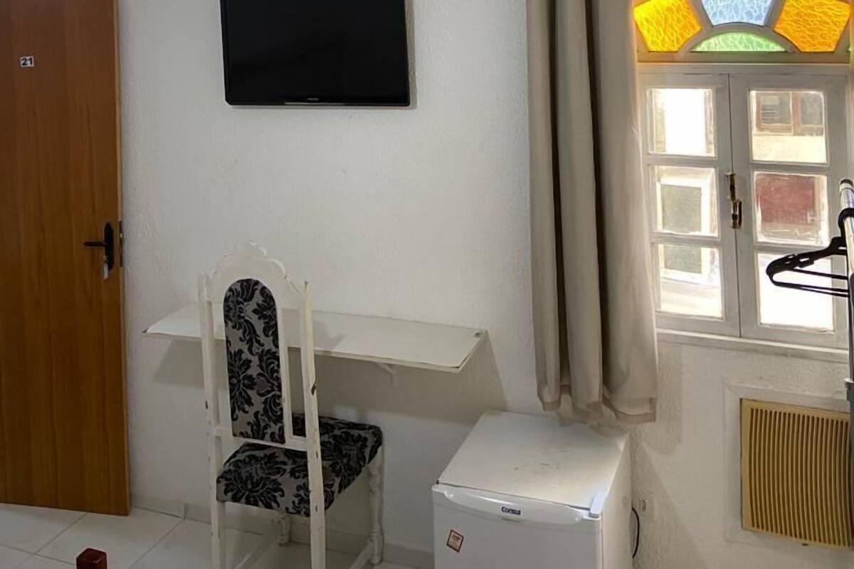 Una habitación con un escritorio blanco, una silla, un televisor montado en la pared, un ventilador de techo, una ventana con cortinas y un radiador.