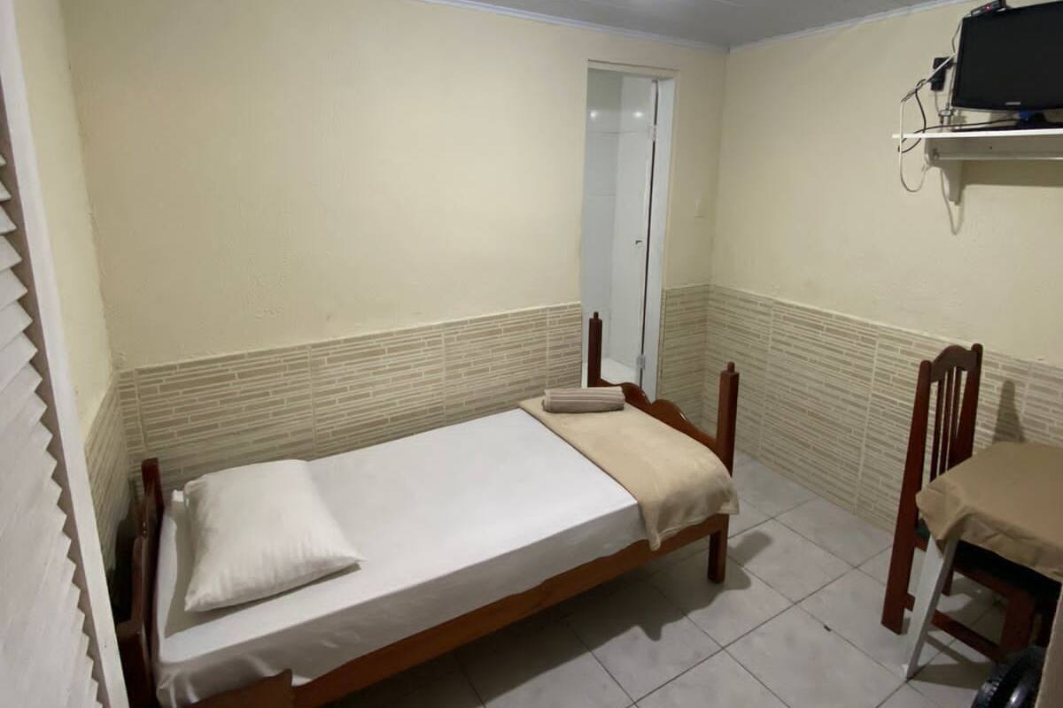 Una habitación pequeña con cama individual, estructura de cama de madera, mesita de noche, una silla y televisor montado en la pared.