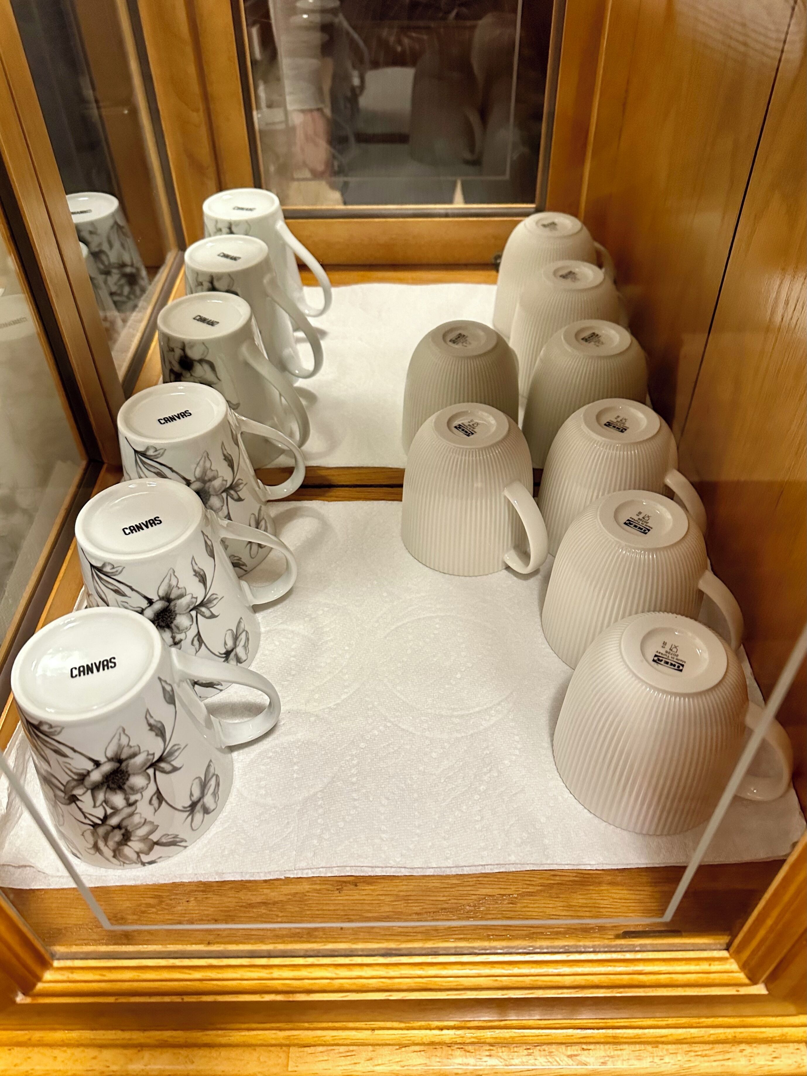 Diverses tasses de ceràmica amb motius florals i la paraula "CANVAS" escrites, organitzades a una estant dins un armari de fusta.